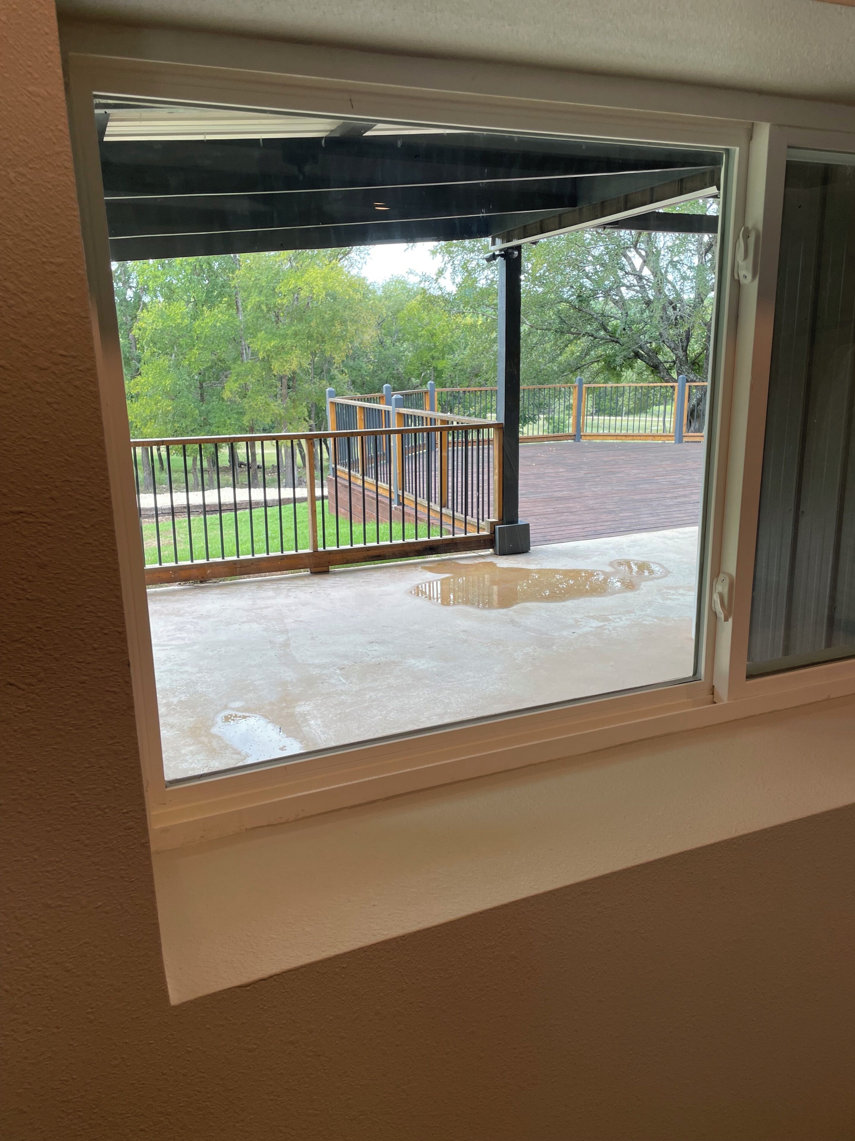 Una ventana con vista a una terraza de madera, baranda metálica y árboles verdes.