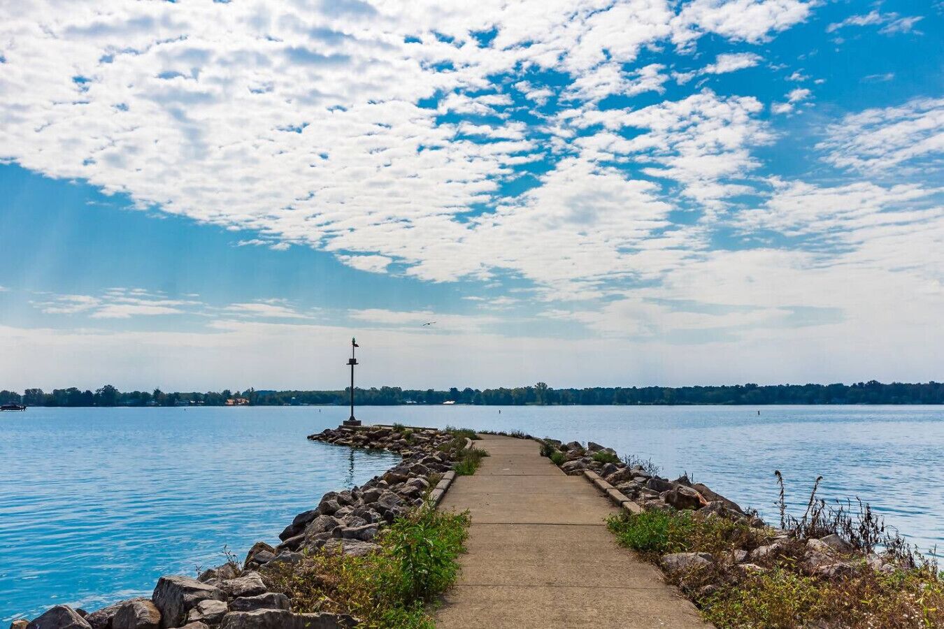 Un chemin de béton mène à une jetée rocheuse qui s’avance dans un lac paisible, sous un ciel dégagé.