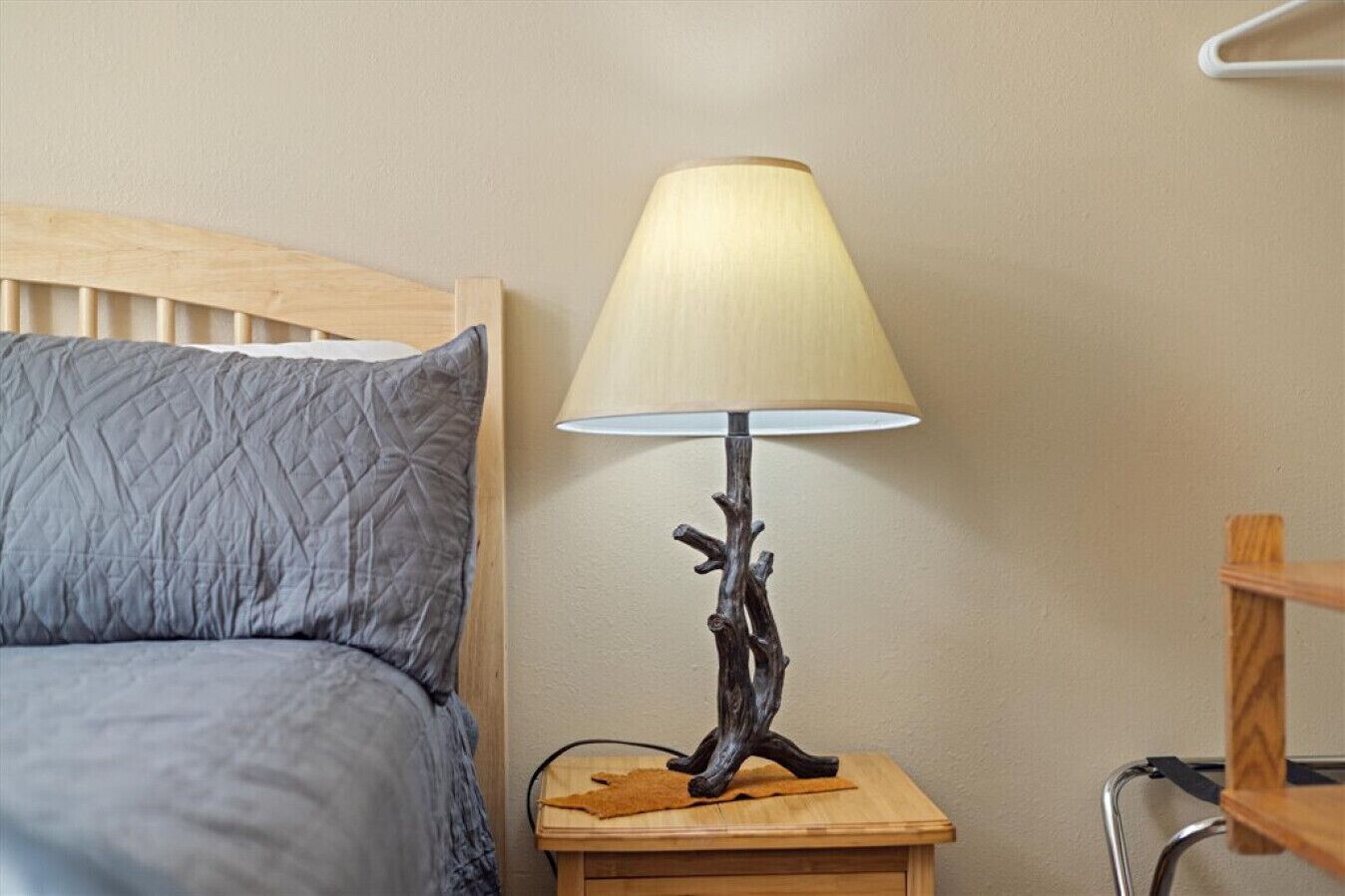 A bed with a quilted gray blanket and a wooden headboard, a bedside table with a lamp featuring a wooden base and a beige lampshade, and a metal chair with a wooden seat.