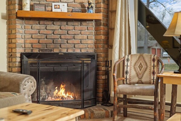 A cozy living room with a brick fireplace, a wooden shelf, a patterned armchair, and a table with a remote control. - Gunslinger Gateway Red Lodge (Red Lodge)