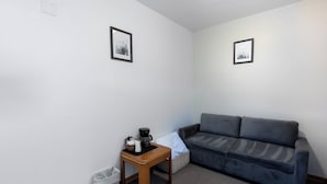 Une petite pièce moderne, avec un canapé gris, une table d’appoint en bois sur laquelle se trouvent une machine à café et un panier, et deux tableaux encadrés accrochés aux murs.