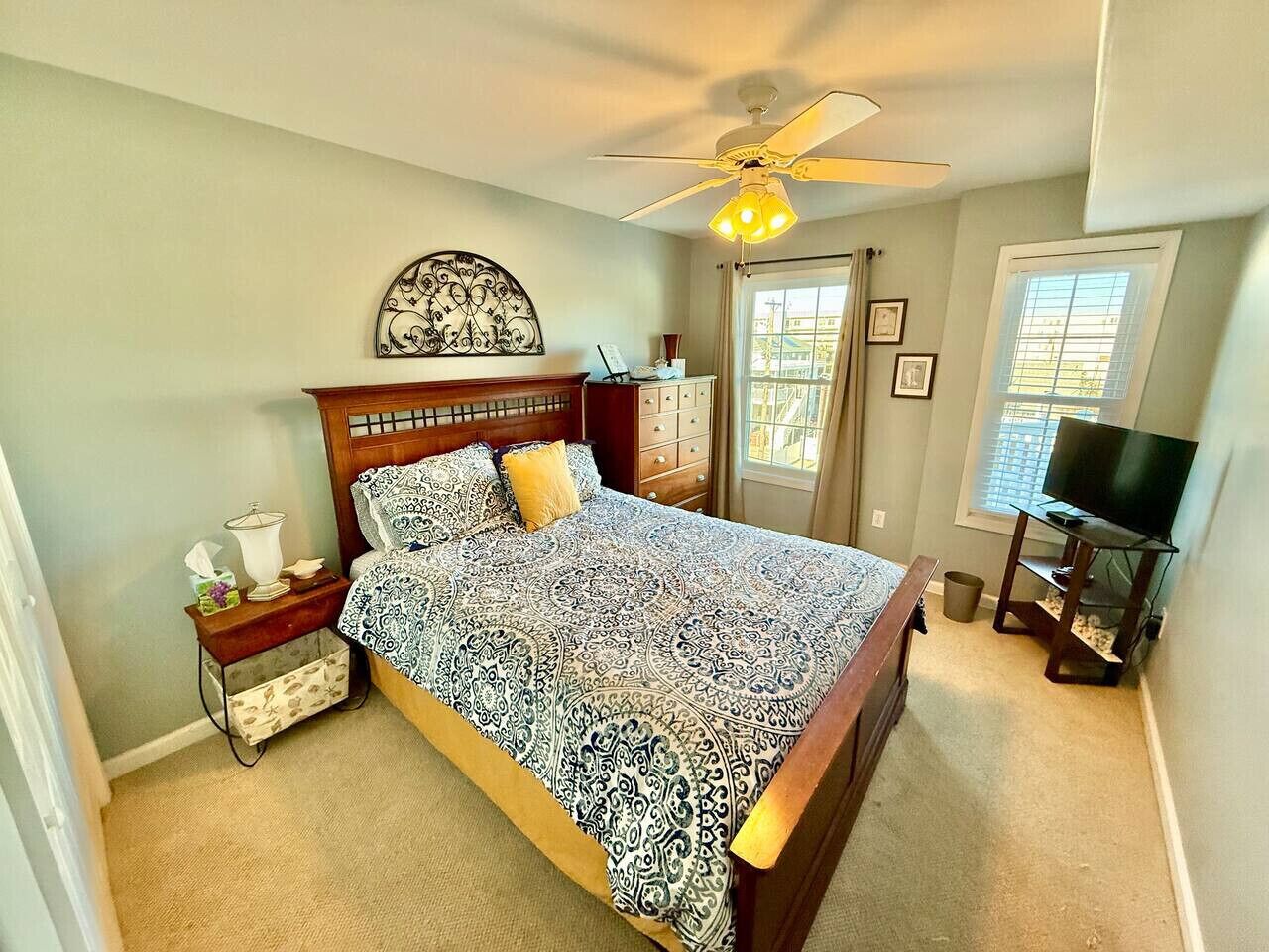 A bedroom with a bed, nightstand, dresser, ceiling fan, TV, and window with curtains.