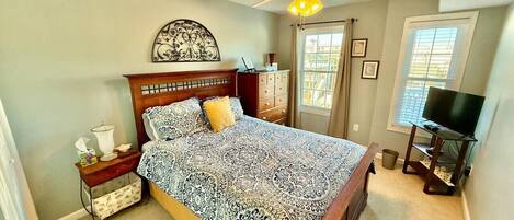 A bedroom with a bed, nightstand, dresser, ceiling fan, TV, and window with curtains.