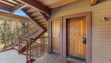 A wooden staircase leading to a balcony with a metal railing, a wooden door with a doormat, and a view of trees.