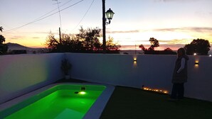 Une piscine éclairée d’une lumière verte, entourée d’un mur blanc orné de lumières, et un coucher de soleil en arrière-plan.