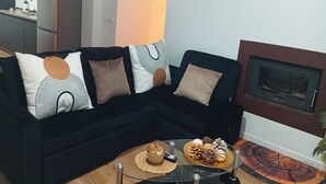 Un salon moderne avec un canapé de couleur foncée, des coussins décoratifs, une table basse en verre sur laquelle se trouve un plateau de chocolats, et une cheminée.