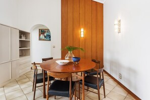 A dining area with a wooden table, chairs, a vase with a plant, and a wall-mounted light fixture. - Commodore 8 - Top Floor Views (Noosa Heads)