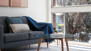 A modern living room with a grey sofa, a glass coffee table, and a large window offering a view of trees and a brick wall. - Prickly Pear Stays in Merewether - Ridge St (Merewether)