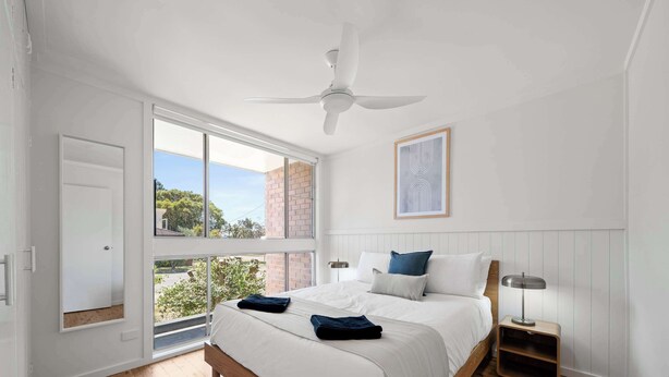 A bedroom with a large window, a ceiling fan, a bed with pillows, and a bedside table with a lamp. - Prickly Pear Stays in Merewether - Ridge St (Merewether)