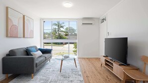 A modern living room with a grey sofa, a glass coffee table, a wooden TV stand, and a large TV. - Prickly Pear Stays in Merewether - Ridge St (Merewether)