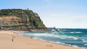 A beach with a rocky cliff, a large ship in the distance, and a parking area with cars. - Prickly Pear Stays in Merewether - Ridge St (Merewether)