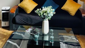 Ein Wohnzimmer mit einem schwarzen Sofa, zwei gelben Kissen, einer weißen Vase mit Blumen, einer runden Wanduhr und einem Glastisch.