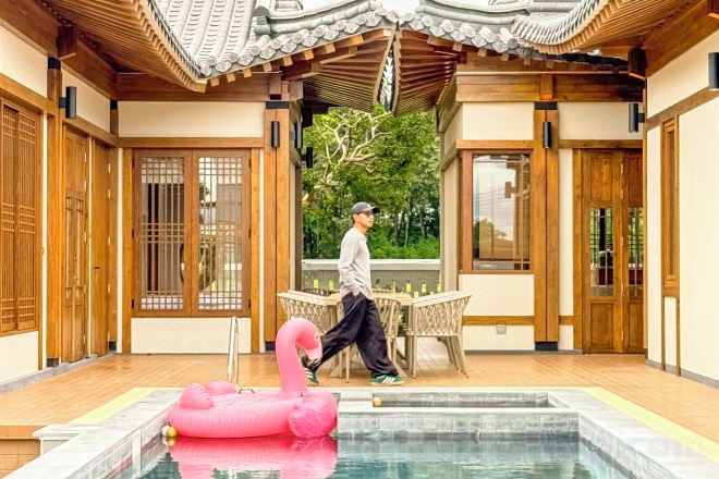 Un bâtiment de style japonais traditionnel, avec un intérieur en bois, une piscine agrémentée d’un flotteur en forme de flamant rose, et une personne qui se promène.