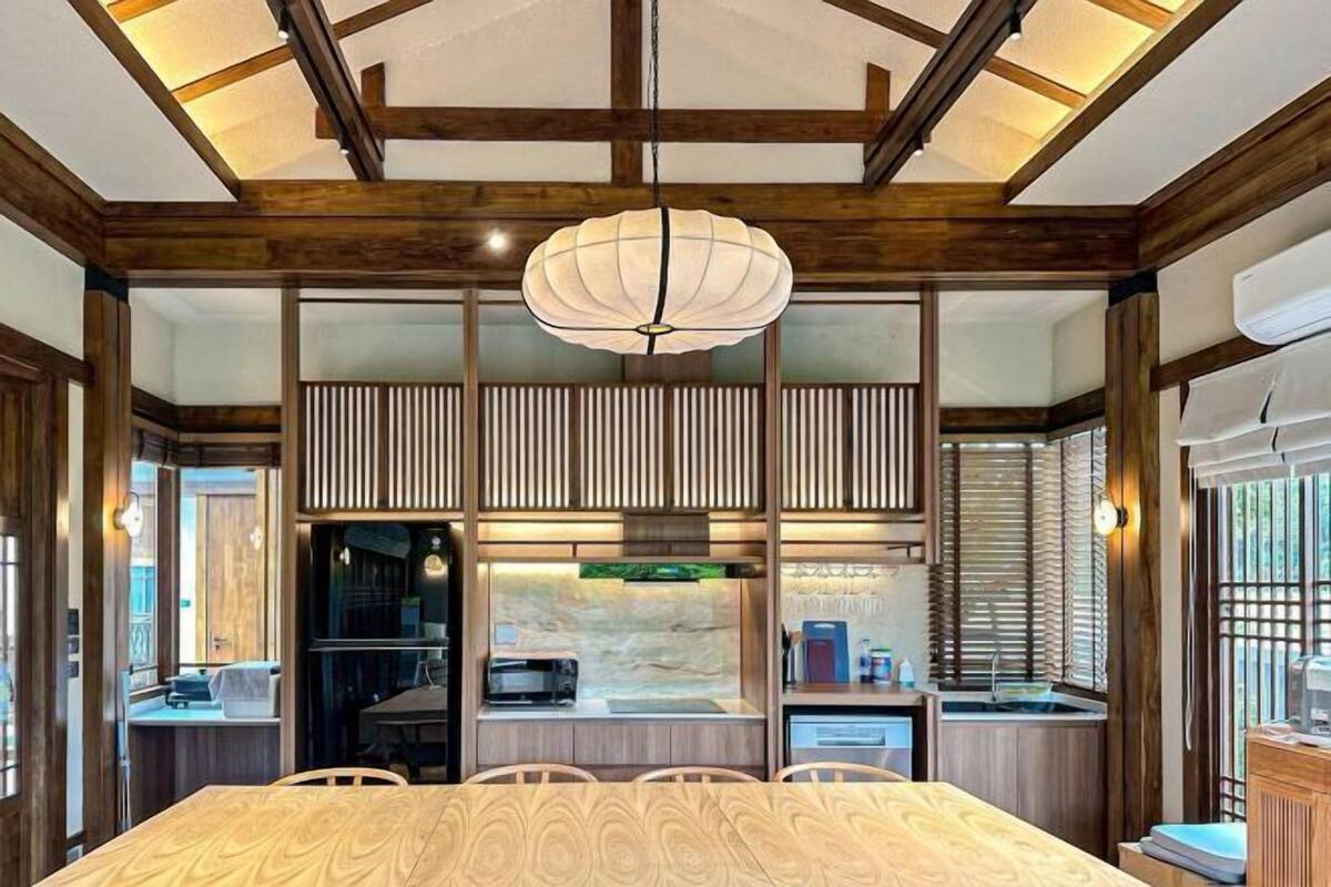 Une pièce de style japonais traditionnel, avec une table à manger en bois, un luminaire suspendu et un coin cuisine visible à travers une porte coulissante.