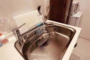 Un lavamanos de acero inoxidable con un escurreplatos, una tabla de cortar y un microondas al fondo.