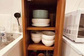Une cuisine équipée d’un four à micro-ondes, d’une étagère en bois où sont empilés des bols, et d’une cuillère posée sur le comptoir.