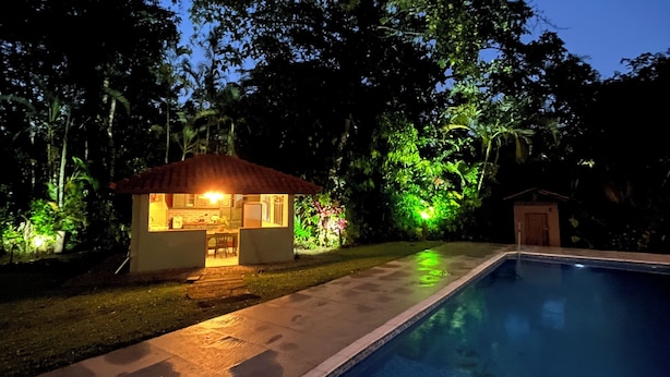 A swimming pool with a tiled deck and a small house with a red roof and a lit interior. - House with pool and 4 suites in Bertioga (Bertioga)