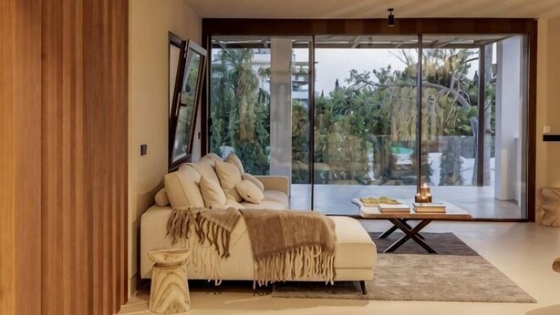 A modern living room with a sofa, a coffee table, and a large glass door leading to an outdoor area. - All About Santa Ana Villa (Marbella)