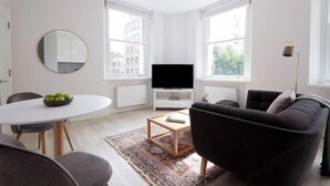 A modern living room with a flat-screen TV, a round mirror, a dining table with chairs, a black sofa, and a patterned rug. - 4VH Virginia House, 31 Bloomsbury Way by City Living London (London)