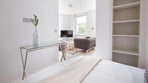 A modern bedroom with a glass desk, a bed with a white headboard, a grey sofa, and built-in shelving. - 4VH Virginia House, 31 Bloomsbury Way by City Living London (London)