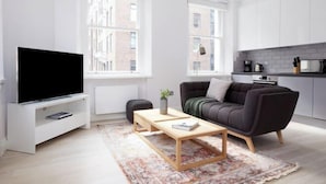 A modern living room with a flat-screen TV, a sofa, a coffee table, and a kitchen area with cabinets and appliances. - 4VH Virginia House, 31 Bloomsbury Way by City Living London (London)