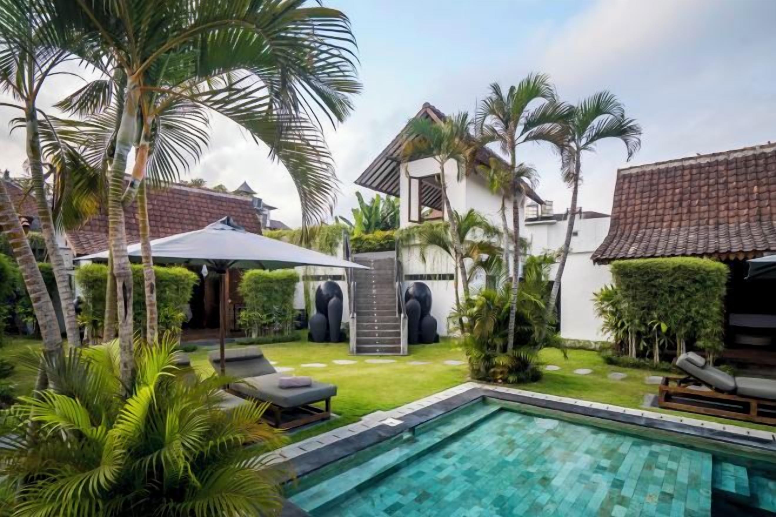 A swimming pool with a sun lounger and umbrella, surrounded by palm trees and a well-maintained lawn.
