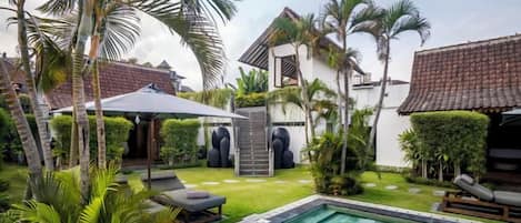 A swimming pool with a sun lounger and umbrella, surrounded by palm trees and a well-maintained lawn.