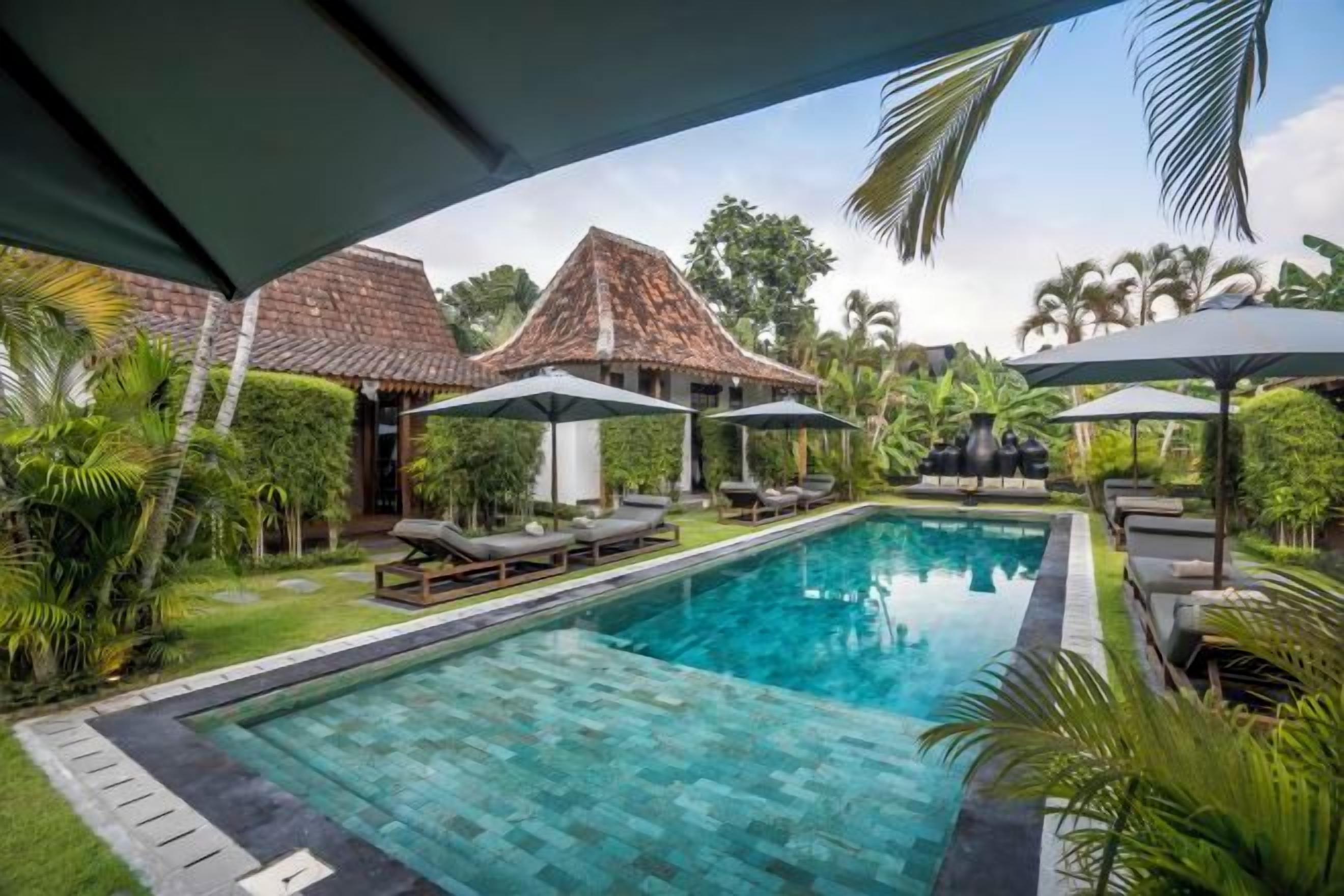 Una piscina con sillones reclinables y sombrillas, rodeada de vegetación y edificios tradicionales.
