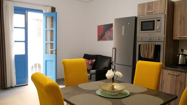 Een moderne keuken met een inbouwoven, magnetron en koelkast. Er staat een eettafel met gele stoelen bij, en een vaas met bloemen. In de achtergrond is een blauwe deur met glaspanelen te zien.
