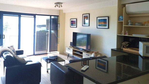 A modern living room with a flat-screen TV, a sofa, a coffee table, and a kitchen area with a microwave and a small refrigerator. - Sails Unit 25 (Sunshine Coast)