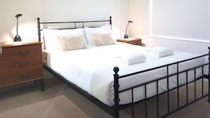 A bedroom with a metal bed frame, white bedding, a wooden nightstand with drawers, and a bedside table with a lamp. - Sails Unit 25 (Sunshine Coast)