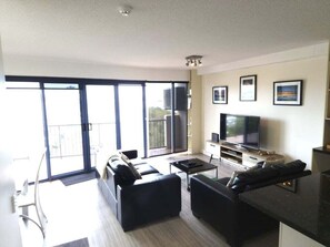 Een moderne woonkamer met een flatscreen televisie, zwarte banken, een salontafel en een balkon met grote ramen.