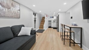 A modern living room with a grey sofa, a small table, and a staircase leading to a kitchen area. - The Millertime Den (Brantford)