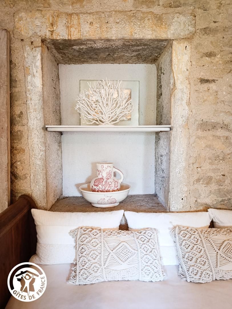 A rustic interior with a stone wall, a shelf with a decorative piece and a ceramic pitcher, and a cushioned bench with patterned pillows.