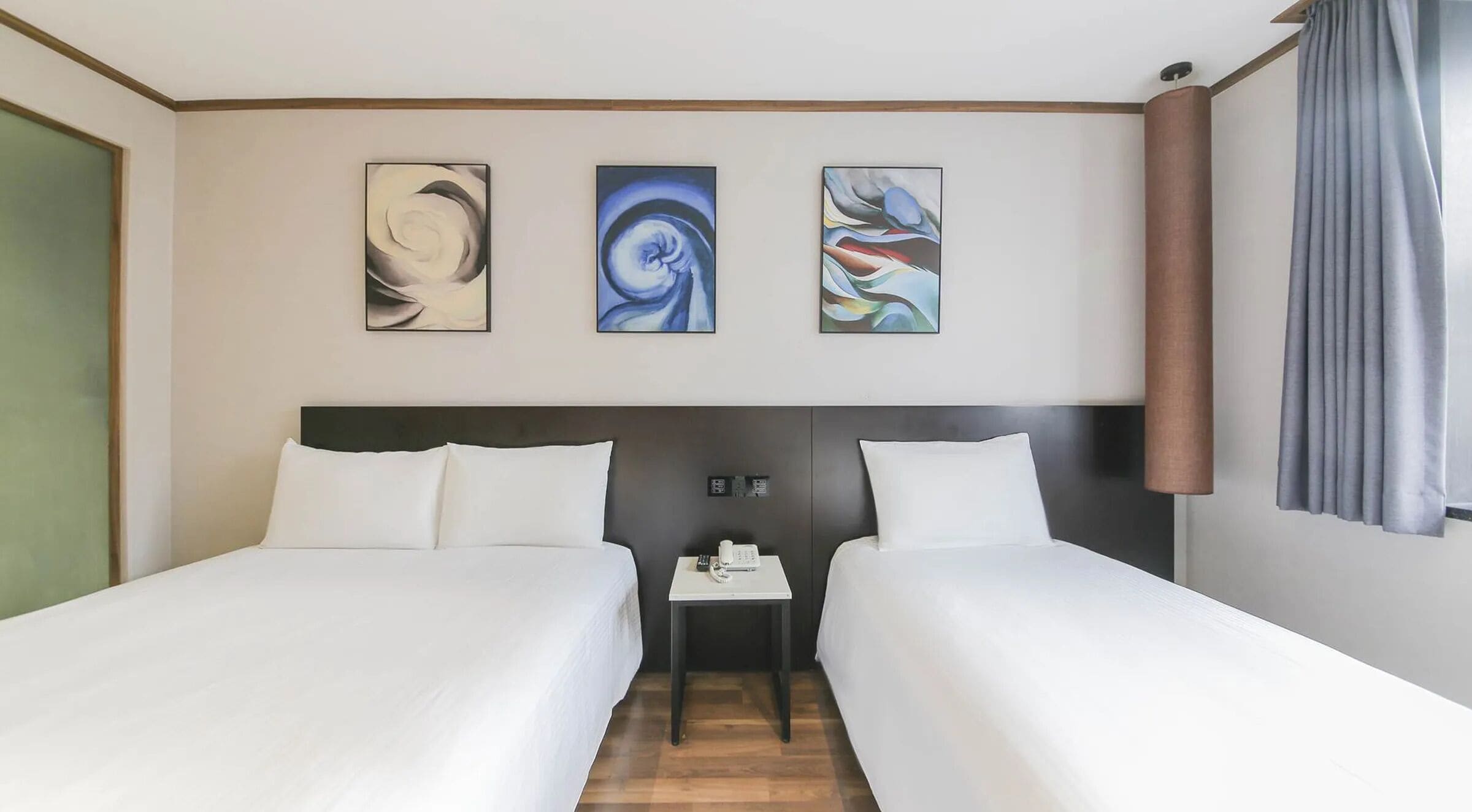 A hotel room with two beds, a small table with a phone, and three abstract paintings on the wall.