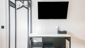 Habitación de hotel con un escritorio blanco, una silla negra, un televisor montado en la pared y una mesita con un dispensador de pañuelos de papel y un atomizador.