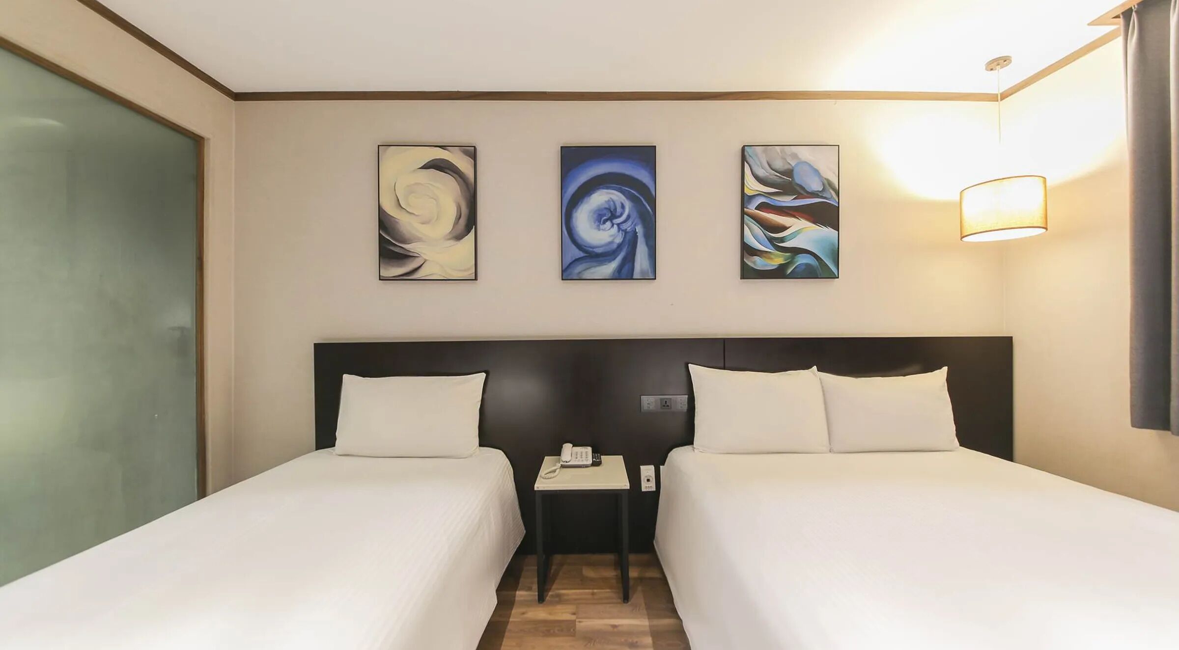 A hotel room with two beds, a small bedside table with a phone, and three abstract paintings on the wall.