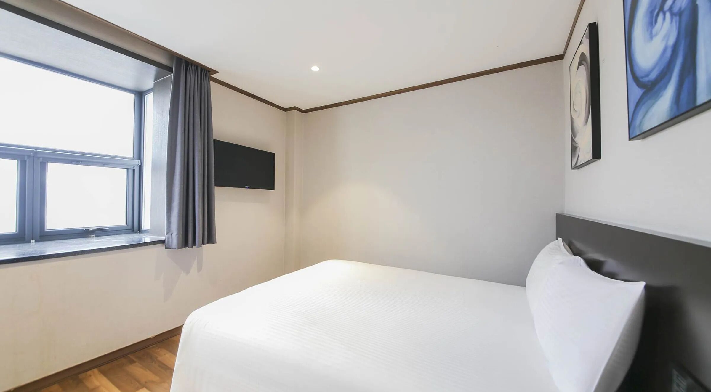 Une chambre d’hôtel avec un lit, une fenêtre avec des rideaux, un téléviseur fixé au mur et un tableau accroché au mur.