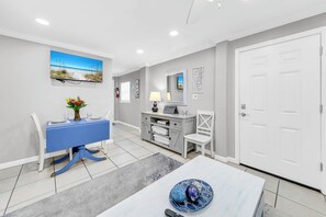 A modern living room with a blue dining table, white chairs, a grey sofa, a grey cabinet, and a white door. - 🦩Book Me Baby! Swords Beach House in Panama City Beach~Golf Cart Included🦩 (Panama City Beach)