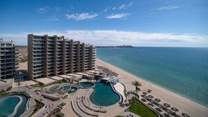 Een hoog hotel met meerdere balkons met uitzicht op een strand en een zwembadgedeelte met ligbedden en parasols.