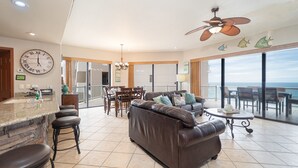 Een ruime woonkamer met een leren bank, eettafel en stoelen. Een grote klok hangt aan de muur, en er is een balkon met uitzicht op de oceaan.