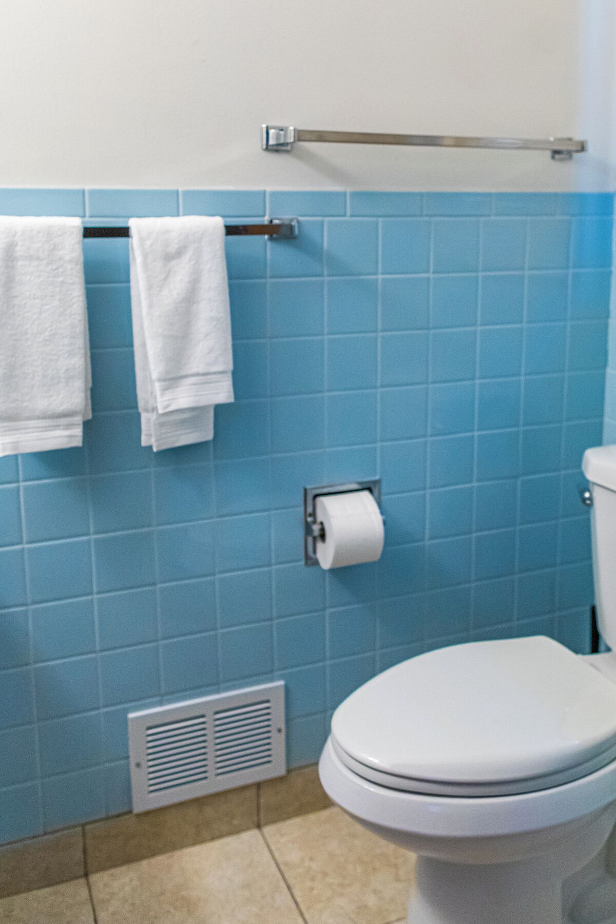 Un baño con azulejos azules, un inodoro, un perchero con dos toallas blancas y un dispensador de papel higiénico.