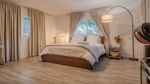 Ein Schlafzimmer mit Holzboden, einem Bett mit weißer Bettdecke, einer Stehlampe und einem Fenster mit Vorhängen.
