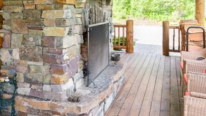 A stone fireplace with a wooden mantel, a wooden deck, wicker furniture, and a view of greenery.