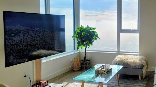 Ein modernes Wohnzimmer mit großem Fernseher, einer Topfpflanze, einem Glastisch und Blick auf die Stadt durch große Fenster.