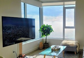 A modern living room with a large TV, a potted plant, a glass coffee table, and a view of the city through large windows. - High Rise Condo in Coquitlam (Coquitlam)