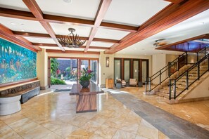 A spacious lobby with a high ceiling, a large mural, a central wooden table with a vase, and a staircase leading to upper floors. - Wailea Beach | Oceanfront 3 BR, Sleeps 6 | Car Incl. w/ 6+ Nights | WBV-PH505 by KBM (Wailea)