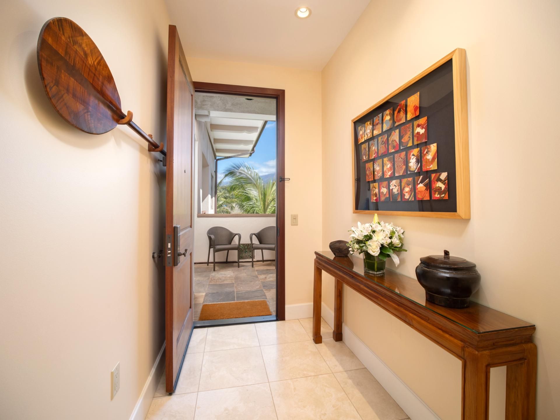 A hallway with a wooden door leading to a balcony with chairs and a view of palm trees.
