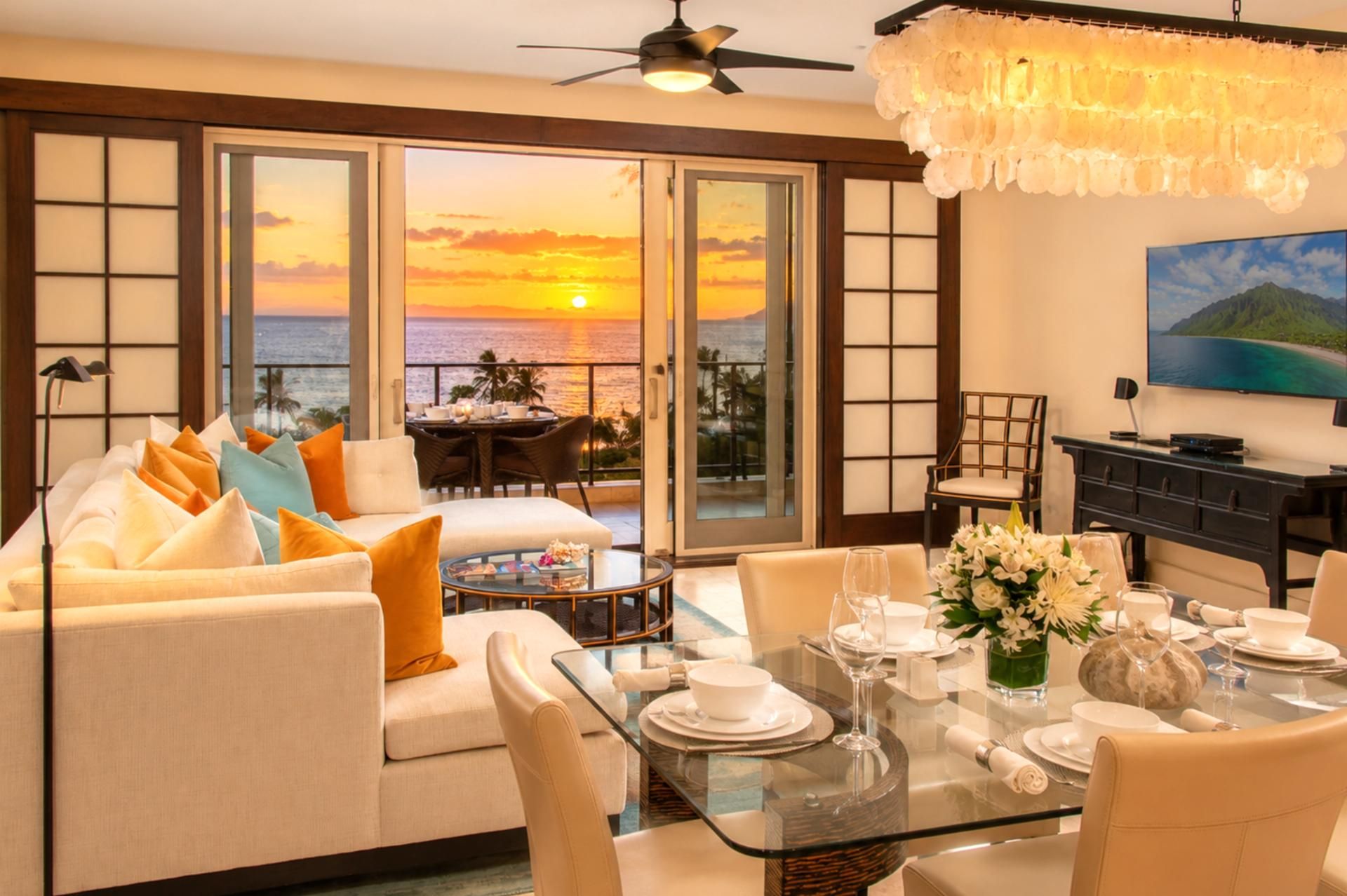 A room with a sofa, dining table, and chairs, with a view of the sunset over the ocean.