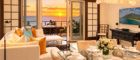 A room with a sofa, dining table, and chairs, with a view of the sunset over the ocean.